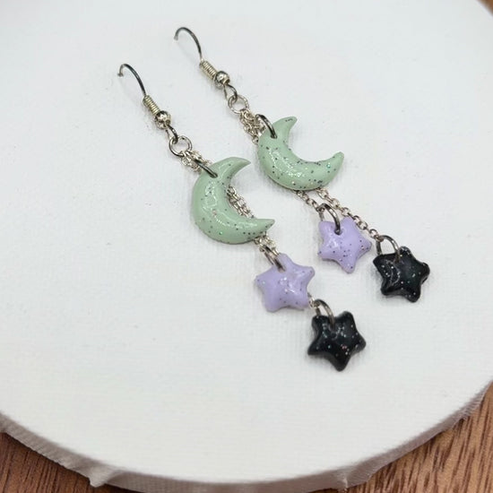 Pair of rainbow glitter green crescent moon and black and purple star earrings with silver hooks on a white background