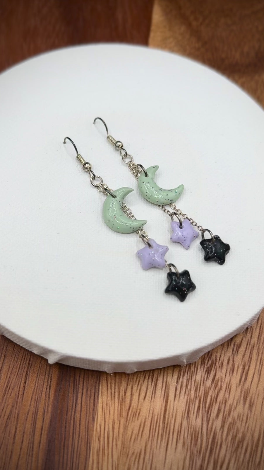 Pair of rainbow glitter green crescent moon and black and purple star earrings with silver hooks on a white background