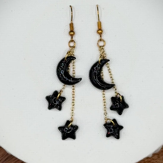 Pair of rainbow glitter black crescent moon and star earrings with gold hooks on a white background