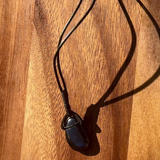 Black hematite stone pendant in a cabochon shape on a leather cord against a wooden background, spinning.
