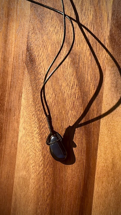 Black hematite stone pendant in a cabochon shape on a leather cord against a wooden background, spinning.