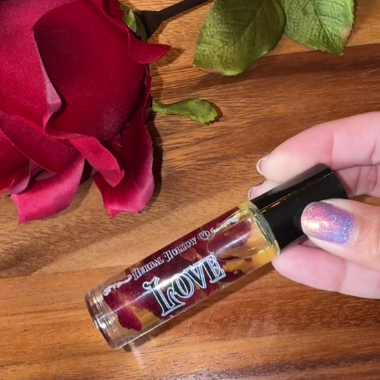Small bottle labeled 'Love' with rose petals suspended in oil inside of it next to a red rose on a wooden surface being held and spun to see the contents and then rolled on the person's wrist.