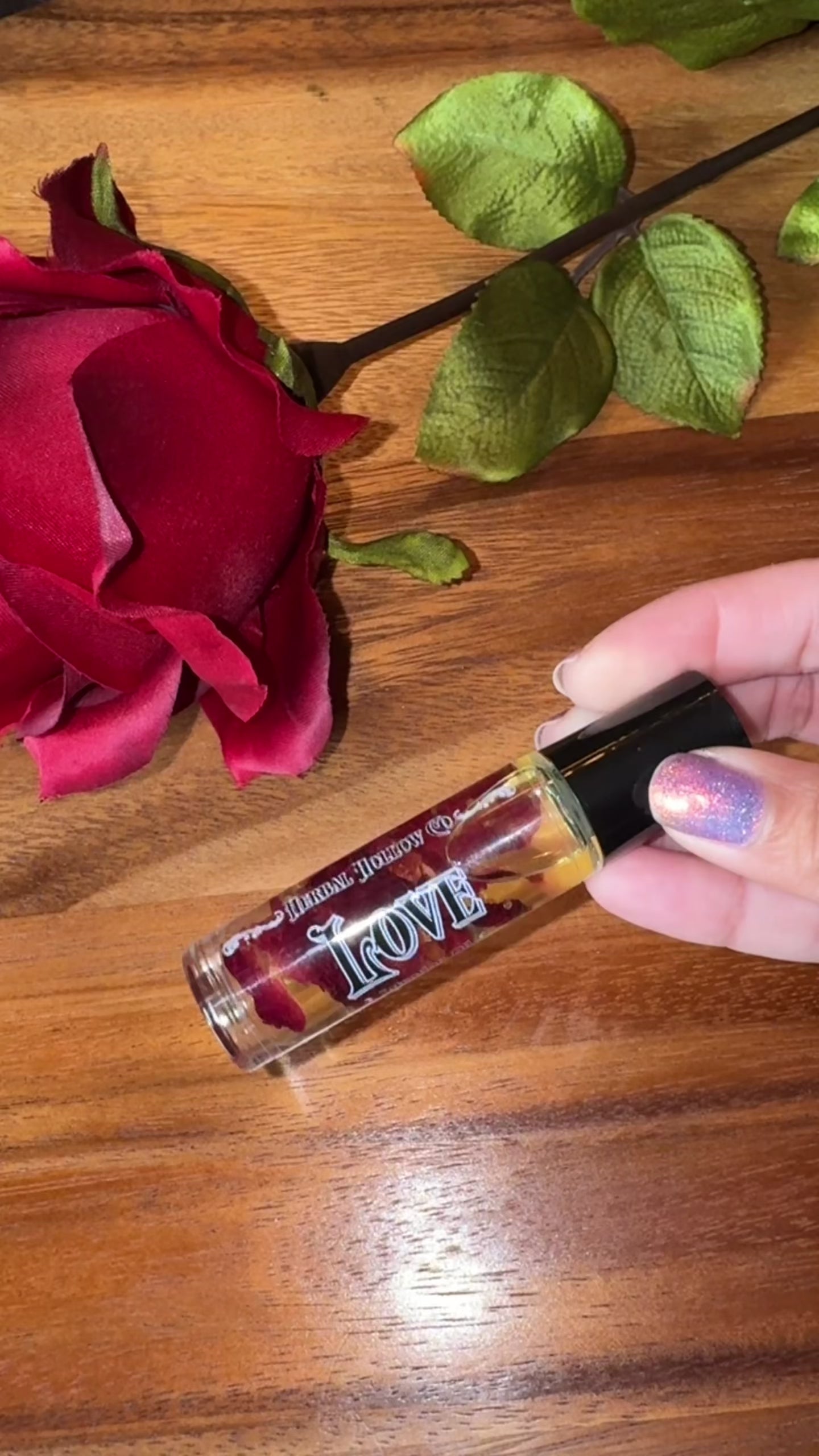 Small bottle labeled 'Love' with rose petals suspended in oil inside of it next to a red rose on a wooden surface being held and spun to see the contents and then rolled on the person's wrist.