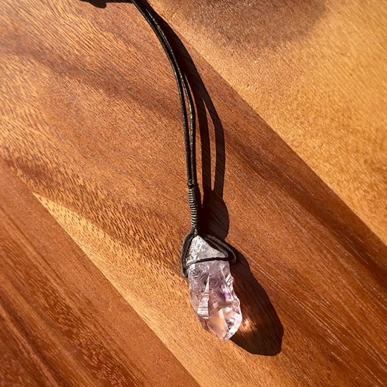 Amethyst stone pendant with inclusions on a leather cord against a wooden background, spinning.