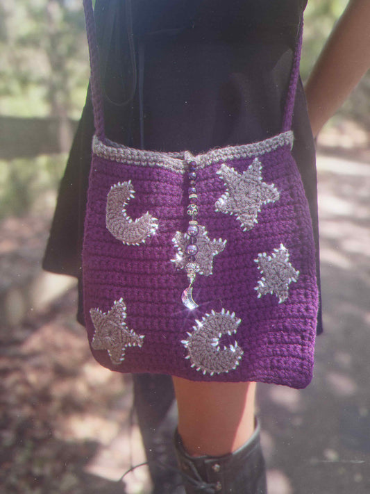 Purple crochet bag with star and moon designs and a moon pendant  held by a person outdoors.