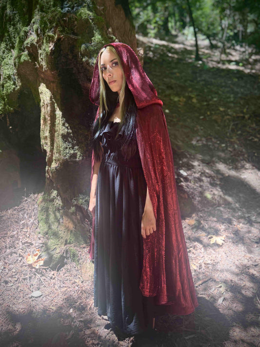 Woman in a dark dress with a red cape with the hood up standing in a forest