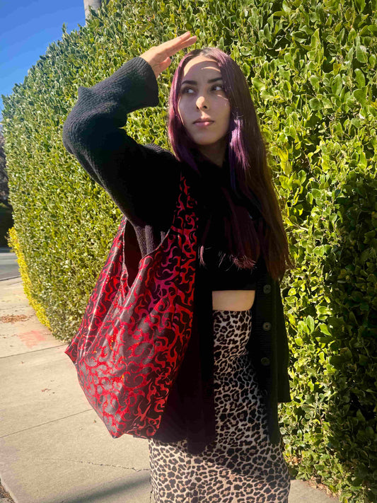 Person wearing a black cardigan, black top, and leopard print skirt with a red flame and black patterned bag, standing against a green hedge.