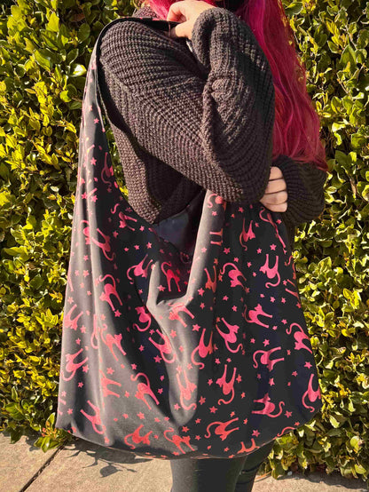 Person with red hair holding a large black with metallic red/pink cats and stars pattern hobo bag against a green hedge background 