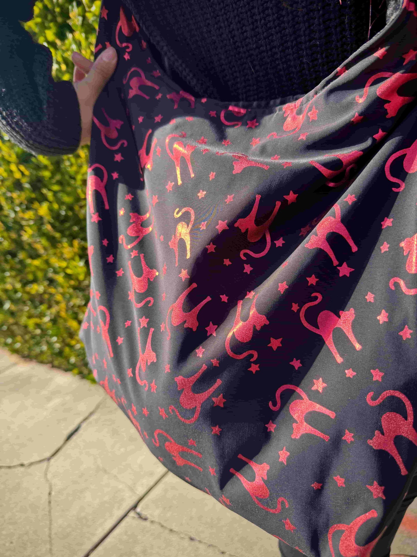 Person holding a large black with metallic red/pink cats and stars pattern hobo bag against a green hedge background 