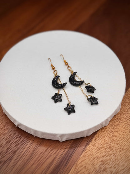 Black moon and star with rainbow glitter earrings and gold hooks on a white circular stand with a wooden background